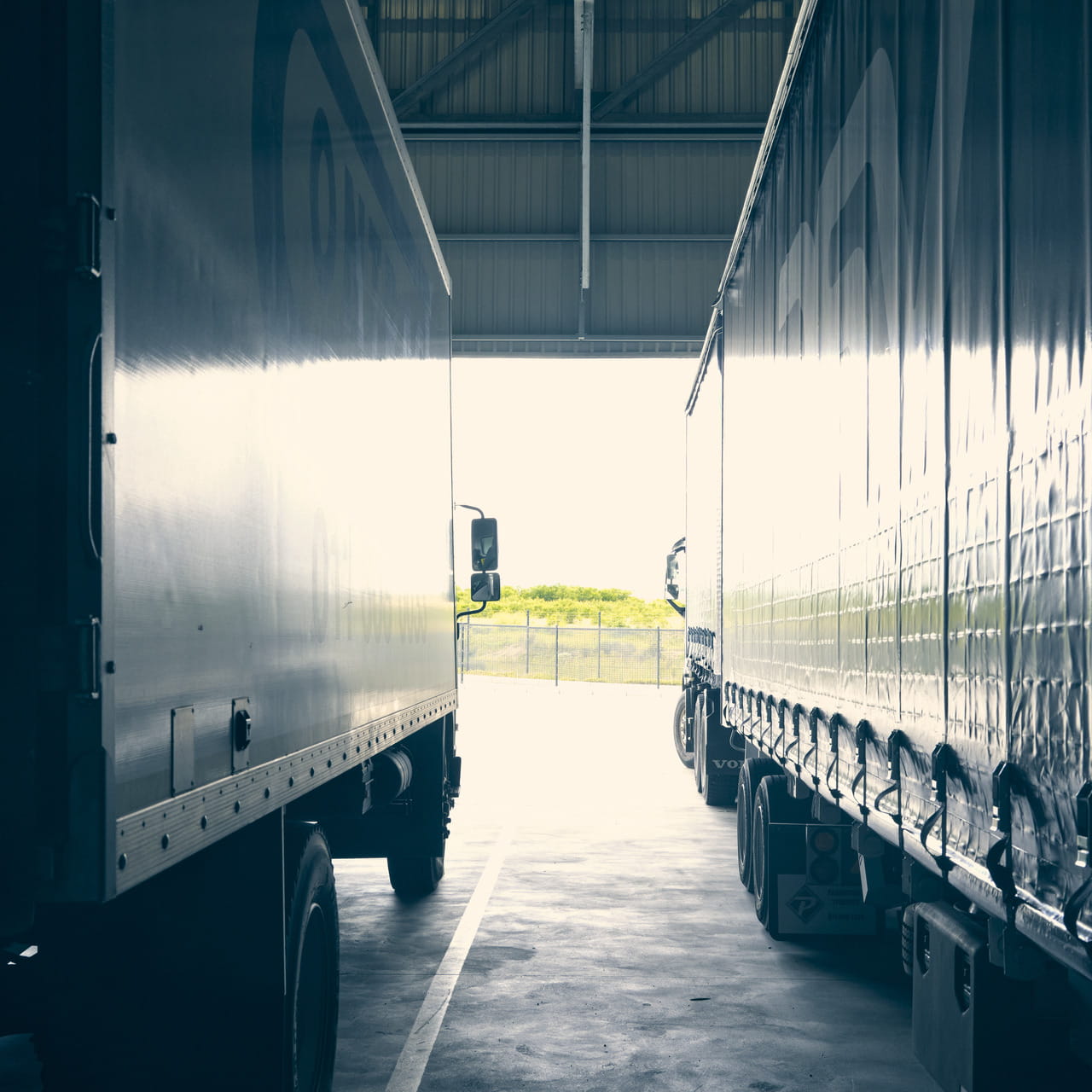 Two semi-trailer trucks parked side by side within a loading dock, under the shelter of logistics facility